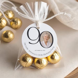 May include: Clear cellophane bag tied with white string, containing gold-wrapped candies. A white tag with the number 90, a photo of a woman, and text that reads "Michelle Johnson's 90th Birthday Party, September 26, 20XX".