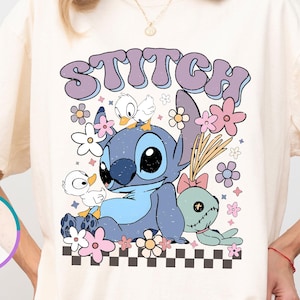 May include: Ivory-colored t-shirt featuring a graphic of Stitch with the word "STITCH" in a retro font. The design includes flowers, ducks, and a small plush toy. The shirt has short sleeves and a relaxed fit.
