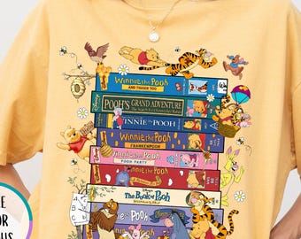 May include: Mustard-colored t-shirt featuring a stack of Winnie the Pooh books with various characters. The shirt has the text "Winnie the Pooh" and "Pooh's Grand Adventure". The shirt also has the text "More Color Options".