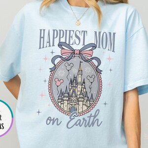 May include: Light blue t-shirt with the text "Happiest Mom on Earth" and a castle graphic. The graphic includes a pink bow, Mickey Mouse balloons, and a castle design. The shirt has short sleeves and a crew neck.