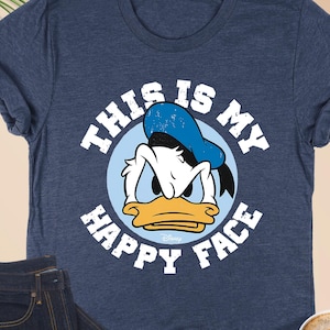 May include: Heather navy blue t-shirt featuring Donald Duck with the text "THIS IS MY HAPPY FACE". The shirt is paired with dark wash jeans, a cup of coffee with a heart design, and tortoiseshell glasses.