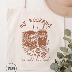 May include: A cream-colored tote bag with the phrase "my weekend is all booked" in brown lettering. The design includes a stack of books, a coffee drink, and floral accents. A beige hat hangs above the bag.