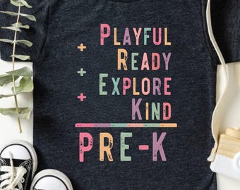 May include: Dark grey heather t-shirt with the words "PLAYFUL READY EXPLORE KIND PRE-K" in colorful text. The shirt is paired with black and white sneakers and a small toy camera.