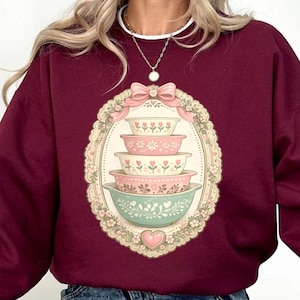 May include: Maroon sweatshirt featuring a stack of vintage-style floral bowls within an oval frame. The bowls are in shades of pink and green, with a decorative bow at the top. The sweatshirt is a solid maroon color.