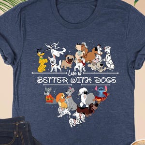 May include: Heather navy blue t-shirt with a graphic of various cartoon dogs arranged in a heart shape. The text "Life is Better With Dogs" is printed below the dogs. A pair of dark blue jeans, a cup of coffee, and glasses are also in the image.