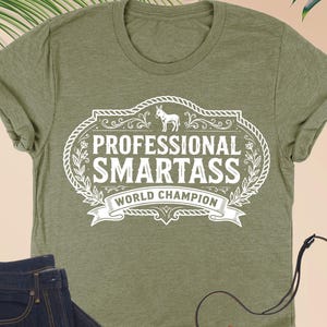 May include: Heather olive green t-shirt with the text "PROFESSIONAL SMARTASS WORLD CHAMPION" in a white decorative frame. The image also includes a pair of dark blue jeans, a brown purse, and eyeglasses.