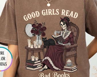 May include: Espresso-colored t-shirt with the text "GOOD GIRLS READ BAD BOOKS". The graphic features a skeleton in a Victorian dress reading a book, with a black cat, flowers, and candles. "MORE COLOR OPTIONS" is also visible.