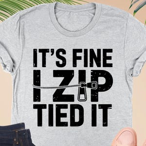 May include: A light gray t-shirt with the text "IT'S FINE I ZIP TIED IT" in black. The image also shows a pair of dark blue jeans, a small pink wallet, and a pair of pink sneakers. The shirt is athletic heather.
