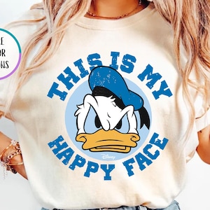 May include: Ivory-colored t-shirt featuring a distressed graphic of Donald Duck with the text "THIS IS MY HAPPY FACE" in blue. The design includes a blue circle around Donald Duck's face and the text. The shirt is a crew neck.