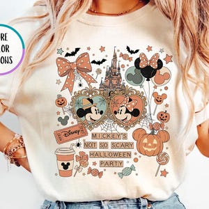 May include: An ivory t-shirt featuring a graphic print with a castle, Mickey and Minnie Mouse in heart-shaped frames, pumpkins, a coffee cup, and candy. The text reads "Mickey's Not So Scary Halloween Party". A circular badge says "More Color Options".