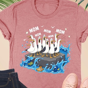 May include: Heather mauve t-shirt with a cartoon seagull graphic. The seagulls are on a rock in the ocean, with the word "MOM" above them. The shirt is paired with dark blue jeans, glasses, and pink sneakers.