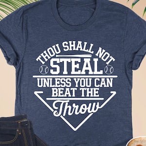 May include: Heather navy blue t-shirt with the text "Thou Shall Not Steal Unless You Can Beat The Throw" in white. The image also includes a pair of dark blue jeans, a cup of coffee, and eyeglasses.