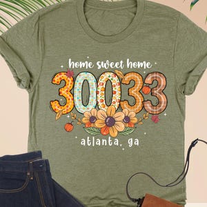 May include: Heather olive green t-shirt with the text "home sweet home 30033 atlanta, ga". The shirt is paired with dark wash jeans, a brown purse, and eyeglasses. The shirt's design includes floral and fall-themed elements.