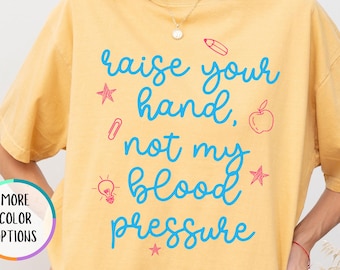 May include: A mustard yellow short-sleeve t-shirt featuring the blue text "raise your hand, not my blood pressure" along with small red graphic icons including stars, a pencil, an apple, and a lightbulb. The shirt has a crew neckline.