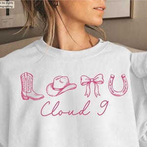 May include: White sweatshirt featuring a pink graphic of a cowboy boot, hat, bow, and horseshoe, with the text "Cloud 9" below. A casual, comfortable top, perfect for everyday wear. The design has a country theme.
