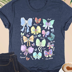 May include: Heather navy blue t-shirt with a colourful bow design and princess names. The bows are in various colours and styles, with names like Ariel, Cinderella, and Snow White. A pair of dark blue jeans and a cup of coffee are also in the image.