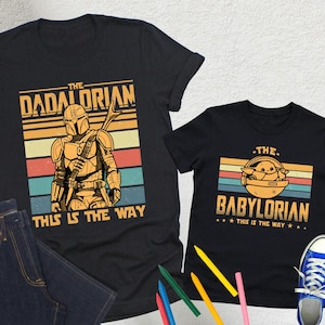 Puede incluir: Dos camisetas negras con dise&ntilde;os de Star Wars. Una dice "The Dadalorian" con una ilustraci&oacute;n de Mandaloriano, la otra "The Babylorian" con Grogu. Ambas camisetas muestran "This is the Way". La imagen tambi&eacute;n incluye unos vaqueros azules y zapatillas azules.