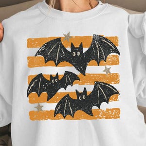 May include: A white crewneck sweatshirt featuring a graphic print of three black bats flying over four horizontal orange distressed stripes. Small grey stars are interspersed between the bats and the orange stripes on the front of the garment.