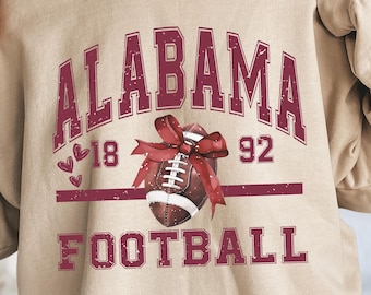 May include: Sand-colored sweatshirt with the word "ALABAMA" in maroon block letters. Below the word "ALABAMA" is a football with a maroon bow, the numbers "18" and "92", and the word "FOOTBALL".