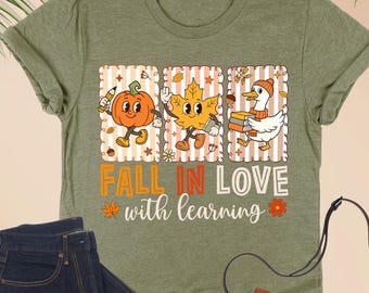 May include: Heather olive green t-shirt with a fall-themed graphic. The graphic features a pumpkin, leaf, and duck with books, and the text "FALL IN LOVE with learning". Dark blue jeans, a brown purse, and glasses are also in the image.