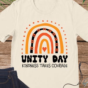 May include: Cream-colored t-shirt with a rainbow graphic in orange, black, and red, and the words "UNITY DAY KINDNESS TAKES COURAGE" in black. The shirt is paired with blue jeans and a brown purse.