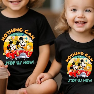 May include: Two children wearing black t-shirts with a colorful graphic of Mickey Mouse and friends in a red car. The text on the shirt reads "NOTHING CAN STOP US NOW!" The shirts are short-sleeved.