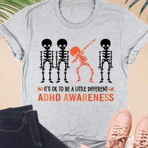 May include: Athletic heather t-shirt with a skeleton graphic and the text "IT'S OK TO BE A LITTLE DIFFERENT ADHD AWARENESS". The image also includes dark blue jeans, pink sneakers, and a pink wallet.