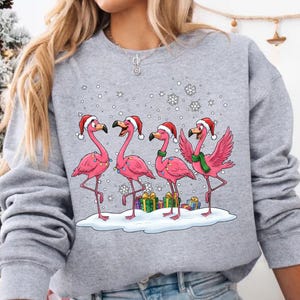 May include: A gray sweatshirt featuring a festive design of four pink flamingos wearing Santa hats and standing in the snow. The flamingos are surrounded by snowflakes and gift boxes, creating a cheerful holiday scene.