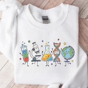 May include: A white long-sleeve sweatshirt featuring a colorful graphic print of cartoon science equipment including a test tube, microscope, flask, DNA strand, and globe, all with smiling faces, surrounded by small multi-colored stars.