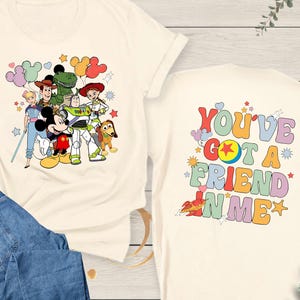 May include: Cream-colored t-shirt featuring a colorful graphic of Disney characters, including Mickey Mouse, Woody, and Buzz Lightyear. The back of the shirt displays the text "YOU'VE GOT A FRIEND IN ME" in a retro font. Paired with blue denim jeans.