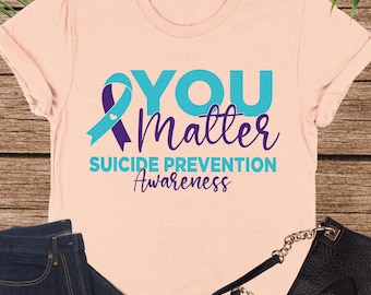 Może przedstawiać: Brzoskwiniowa koszulka z napisem "YOU Matter Suicide Prevention Awareness" w kolorze turkusowym i fioletowym. Na zdjęciu znajdują się również ciemnoniebieskie dżinsy, czarna torebka i czarne baleriny.