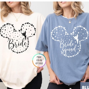 May include: Ivory and blue jean t-shirts with a Mickey Mouse-inspired design. The ivory shirt says "Bride" and the blue jean shirt says "Bride Squad". Both shirts feature a fairy graphic and star and heart accents.