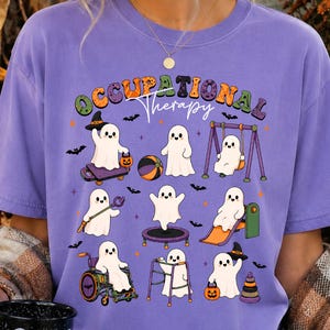May include: A purple t-shirt featuring a graphic of nine white ghosts engaged in various activities, with the text "OCCUPATIONAL Therapy" in colorful, patterned letters. The ghosts are shown using therapy equipment like a wheelchair, walker, and swing.