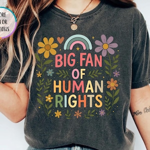 May include: A dark gray t-shirt with the text "BIG FAN OF HUMAN RIGHTS" in colorful letters, surrounded by floral and rainbow graphics. The shirt has a vintage, worn look.