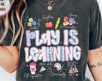 以下が含まれることがあります： 遊び心のあるフォントで「PLAY IS LEARNING」と書かれた濃い灰色のTシャツ。デザインには、学校関連のアイテムや「クリエイティブ」や「シェア」などの言葉のカラフルなイラストが含まれています。シャツには「MORE COLOR OPTIONS」と「PEPPER」の文字も入っています。
