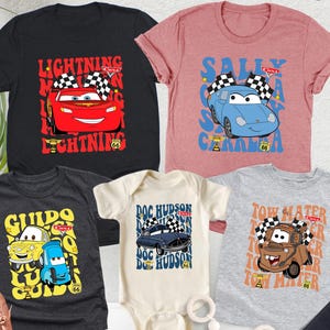 May include: A selection of children's t-shirts and a onesie with cartoon car designs. The shirts are in various colours, such as black, pink, and grey. The designs feature the text "Lightning McQueen", "Sally Carrera", "Guido", "Doc Hudson", and "Tow Mater".