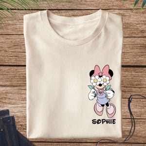 May include: Cream-colored t-shirt featuring a Minnie Mouse graphic with daisy-covered eyes, a pink bow, and a lavender dress. The name "SOPHIE" is printed below the character. The shirt is displayed on a wooden surface with a pair of blue jeans.