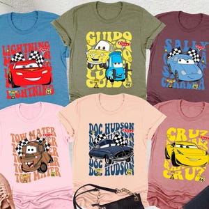 以下が含まれることがあります： アニメ映画「カーズ」のキャラクターが描かれた、様々な色のグラフィックTシャツ。デザインには、ライトニング・マックィーン、メーター、ドック・ハドソン、サリー、グイド、クルーズが含まれます。ピンクのスニーカー、黒いハンドバッグ、ジーンズと一緒に展示されています。