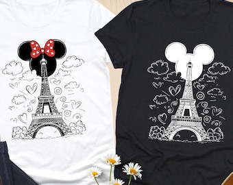May include: Two t-shirts, one white and one black, each with a black and white Eiffel Tower design, Mickey Mouse ears, and hearts. The white shirt has a red bow. A pair of blue jeans and cream-coloured loafers are also visible.
