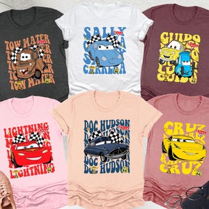 May include: Six t-shirts with cartoon car designs. The shirts are in various colors: dark gray, white, maroon, mauve, peach, and light pink. Each shirt features a different car character with text like 'Tow Mater', 'Sally Carrera', 'Guido', 'Lightning McQueen', 'Doc Hudson', and 'Cruz'.