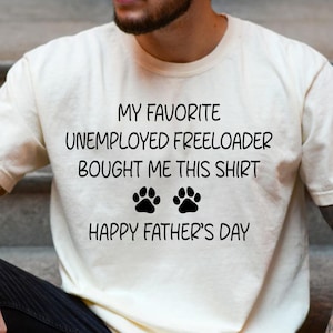 May include: Ivory-colored t-shirt with the text "MY FAVORITE UNEMPLOYED FREELOADER BOUGHT ME THIS SHIRT" and "HAPPY FATHER'S DAY" printed in black. Features two paw prints. A casual, short-sleeved tee.