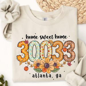 May include: A sand-colored sweatshirt with the text "home sweet home" and the numbers "30033" in a fall-themed design. The sweatshirt also features the text "atlanta, ga" and floral accents.