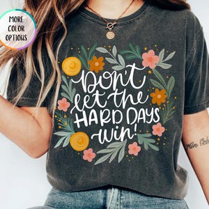 May include: Dark gray t-shirt with the text "Don't let the HARD DAYS win!" in white script, surrounded by a floral wreath of orange and pink flowers and green leaves. The shirt has a vintage, worn look.