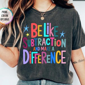 May include: A charcoal gray short-sleeve t-shirt featuring the colorful text BE LIKE SUBTRACTION AND MAKE A DIFFERENCE. The design includes small blue and yellow stars. A circular badge in the upper left corner reads MORE COLOR OPTIONS and a label in the upper right reads PEPPER.