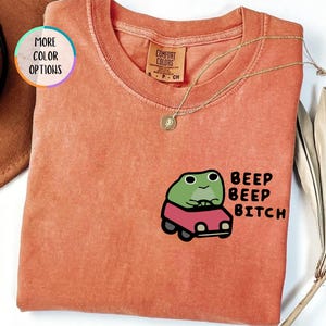 May include: A yam-colored t-shirt with a crew neck and short sleeves. The shirt features a cartoon frog driving a red car, with the text "BEEP BEEP BITCH" printed next to it. A gold necklace is draped on the shirt.