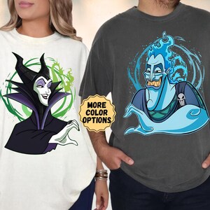 Puede incluir: Dos camisetas de gran tama&ntilde;o, una color crema y otra gris carb&oacute;n. La camiseta crema presenta una imagen de dibujos animados de Mal&eacute;fica, mientras que la camiseta gris muestra a Hades. Ambas camisetas tienen el texto "MORE COLOR OPTIONS" en una pegatina amarilla.