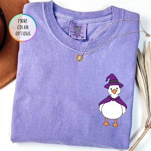 May include: A violet short-sleeve t-shirt with a crew neckline and a graphic of a white duck wearing a purple witch hat and cape. The shirt features a Comfort Colors brand label at the inner neck and a gold-toned necklace resting on the fabric.