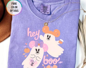 Op de afbeelding: Een violetkleurig T-shirt met korte mouwen en een afbeelding van twee cartoongeesten met muizenoortjes, een pompoen en snoepjes. De tekst luidt "hey boo". Het shirt heeft een ronde hals en een Comfort Colors-label aan de binnenkant van de kraag.