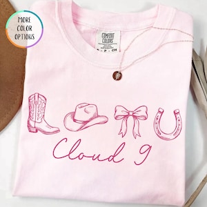 May include: A light pink t-shirt with a crew neck, featuring a cowboy boot, hat, bow, and horseshoe design, with the words "Cloud 9" in a cursive font. A gold necklace with a pendant is draped across the shirt.
