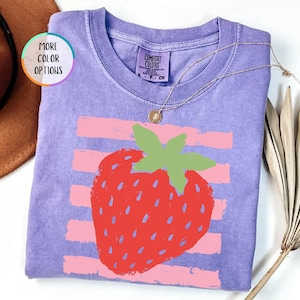 May include: A violet short-sleeved t-shirt featuring a graphic of a red strawberry with green leaves centered over horizontal light pink stripes. The shirt has a crew neckline and a tag that reads Comfort Colors, S, P, CH.